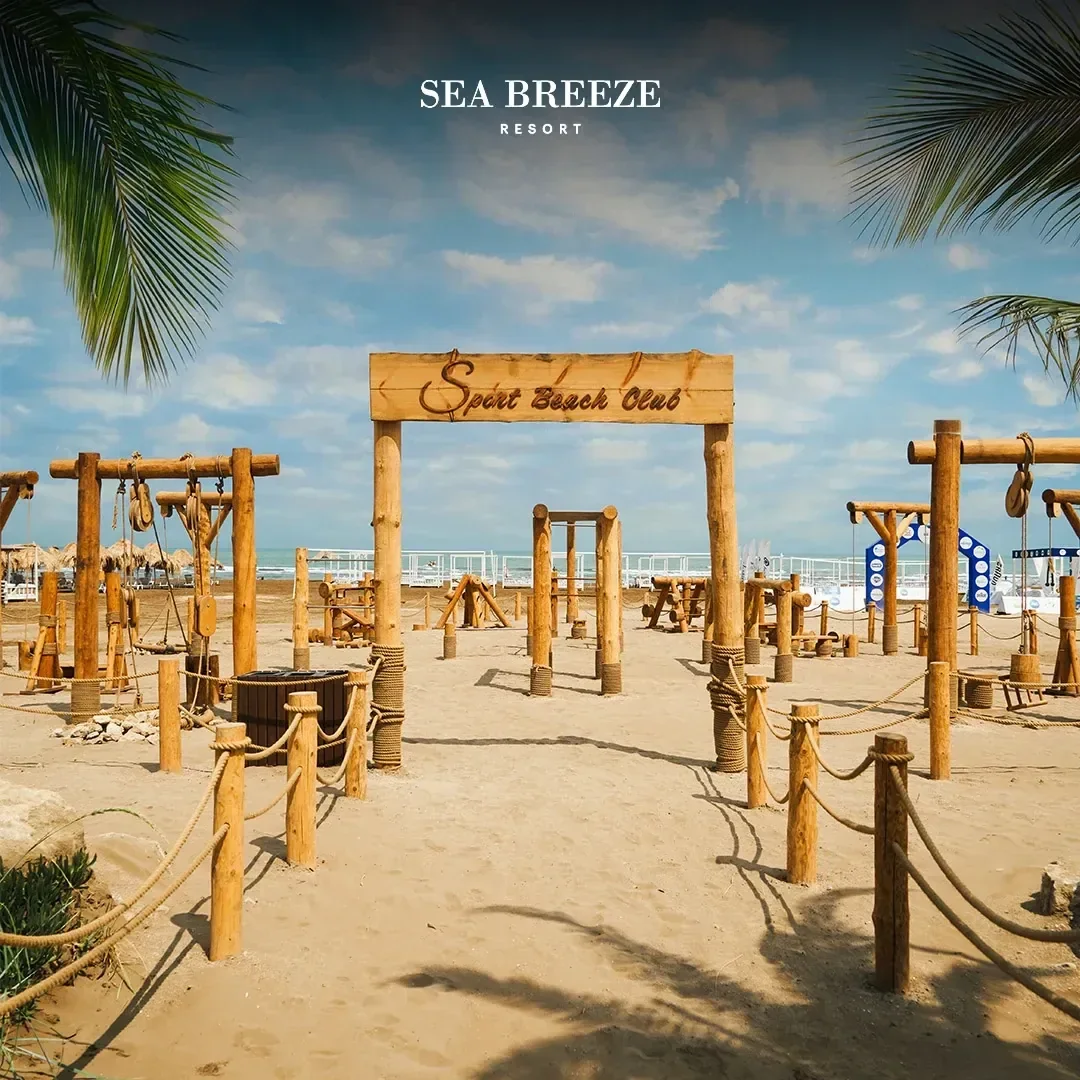Вход в зал: деревянная арка с вывеской Sport Beach Club, канатные ограждения на столбиках и рамы из брёвен на песке