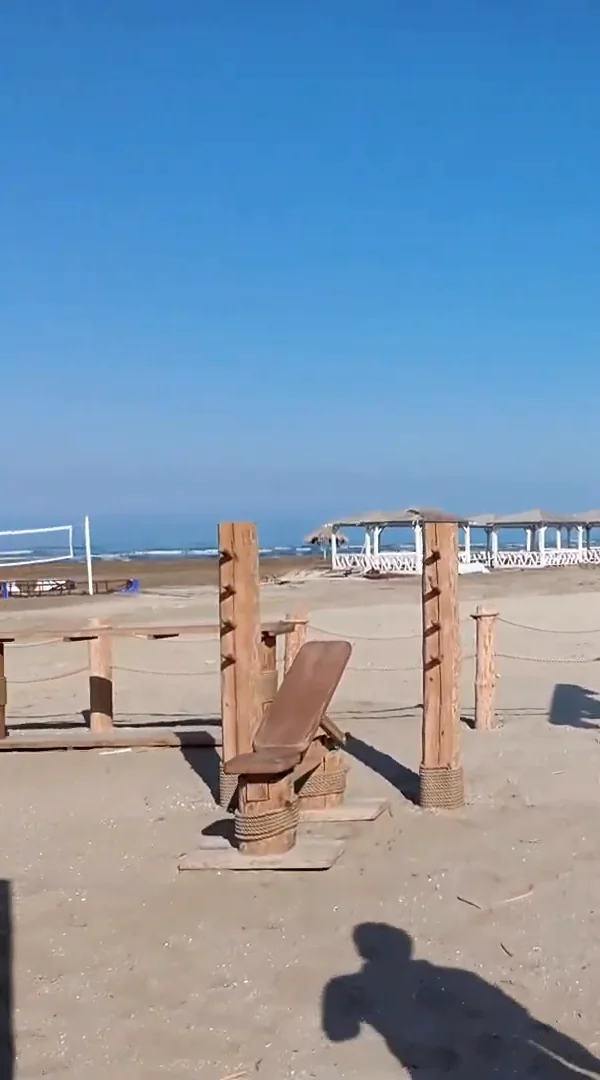 Скамья на чурбаке с наклонной спинкой из доски и столб с колышками; вдали волейбольная сетка